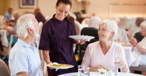 senior living dining room staff serves dinner to an elderly couple